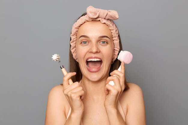 Plan horizontal d'une femme optimiste positive étonnée excitée avec une bande de cheveux debout isolée sur fond gris criant de bonheur en achetant de nouveaux accessoires cosmétiques