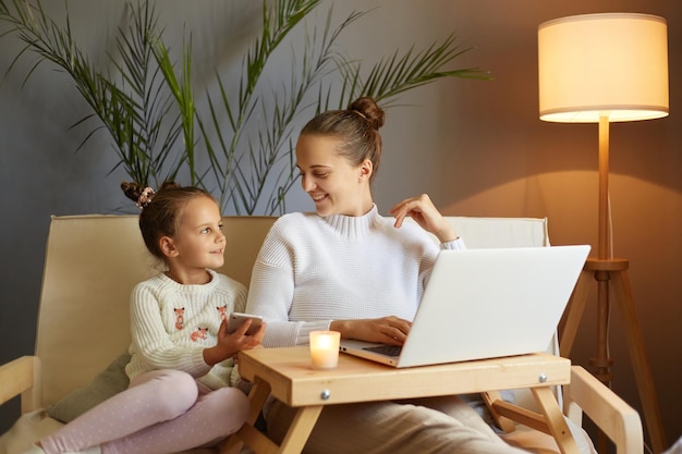 Plan horizontal d'une femme séduisante en pull blanc assise sur un canapé avec sa fille et utilisant un ordinateur portable parlant et profitant du temps ensemble à la maison le week-end