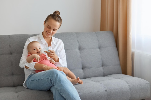 Plan horizontal d'une femme souriante portant une chemise blanche et un jean assis sur un canapé avec une petite fille mignonne enfant buvant de l'eau de la bouteille et assis sur les genoux de la mère