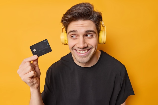 Plan horizontal d'un homme brun heureux titulaire d'une carte de crédit heureux d'obtenir de l'argent sur son compte bancaire porte un casque stéréo sur les oreilles habillé avec désinvolture isolé sur fond jaune pense quoi acheter