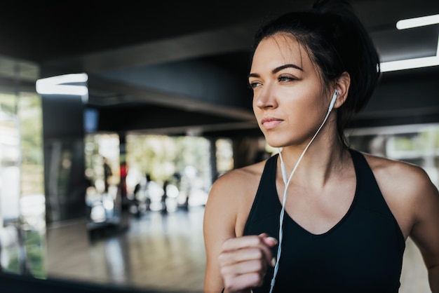 Plan horizontal d'une jeune coureuse de race blanche écoutant de la musique sur des écouteurs tout en courant dans la salle de sport portant des vêtements de sport noirs Concept de sport et de remise en forme