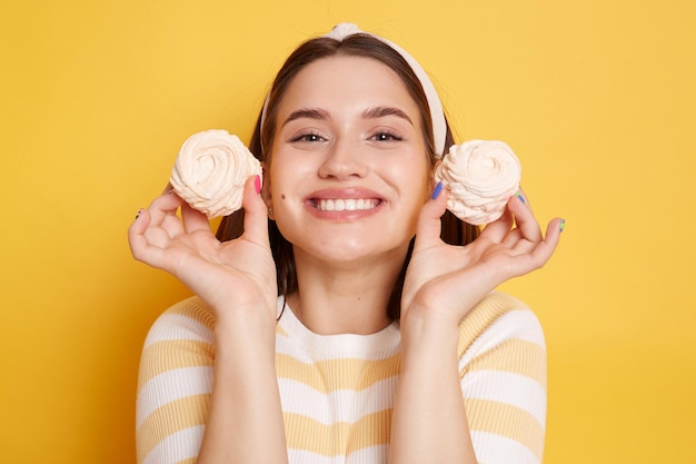 Plan horizontal d'une jeune femme adulte ravie montrant à la caméra de délicieux desserts délicieux sourit largement porte une chemise rayée posant isolé sur fond jaune