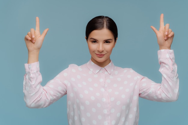 Plan horizontal de la jeune femme brune confiante en chemise formelle pointe vers le haut avec les deux doigts antérieurs