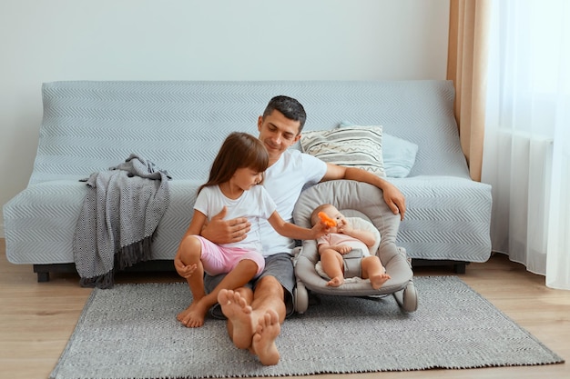 Plan horizontal d'un jeune père optimiste et positif avec ses filles assis par terre près d'un canapé gris, passant du temps avec ses enfants, exprimant son bonheur, étant heureux de jouer avec ses enfants.