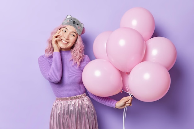 Plan horizontal d'une jolie jeune femme asiatique tenant un tas de ballons gonflés se prépare pour une fête ou une célébration porte une jupe de pull décontractée et un masque de sommeil sur le front isolé sur fond violet