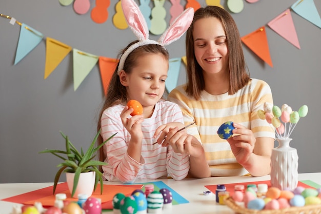 Plan horizontal d'une mère joyeuse optimiste et de sa fille peignant des oeufs de Pâques se préparant pour Pâques assis à table avec une décoration de Pâques contre un mur gris avec des accessoires de fête