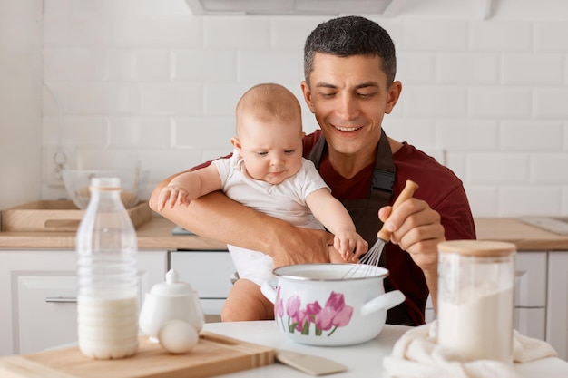 Plan horizontal d'un père satisfait en tablier assis à table avec sa petite fille et cuisinant ensemble dans la cuisine en mélangeant de la pâte pour une pâtisserie maison exprimant le bonheur