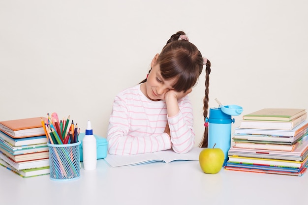 Plan horizontal d'une petite écolière endormie et épuisée aux cheveux noirs et aux tresses assise à table appuyée sur sa main et dormant fatiguée d'étudier toute la journée