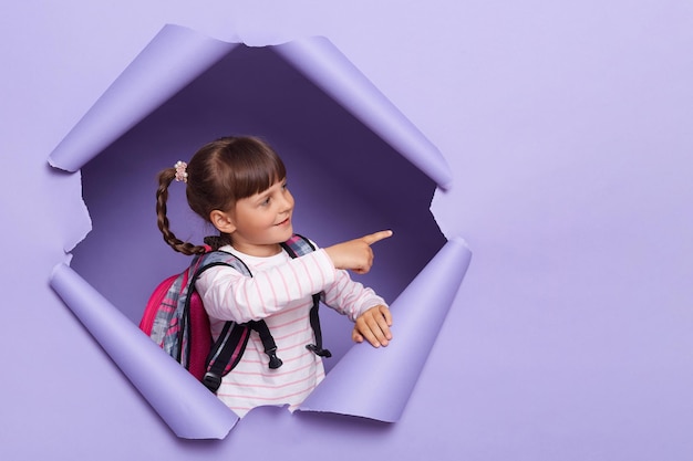 Plan horizontal d'une petite fille aux cheveux noirs avec des tresses portant une chemise rayée posant dans un mur de papier déchiré debout avec un sac à dos et pointant vers l'espace de copie pour la publicité