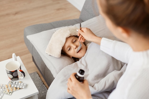 Plan horizontal d'une petite fille malade allongée sur un canapé, d'une mère donnant sa cuillère avec du sirop, d'une famille posant dans une pièce lumineuse, d'un enfant prenant des médicaments contre la toux ou la température élevée.