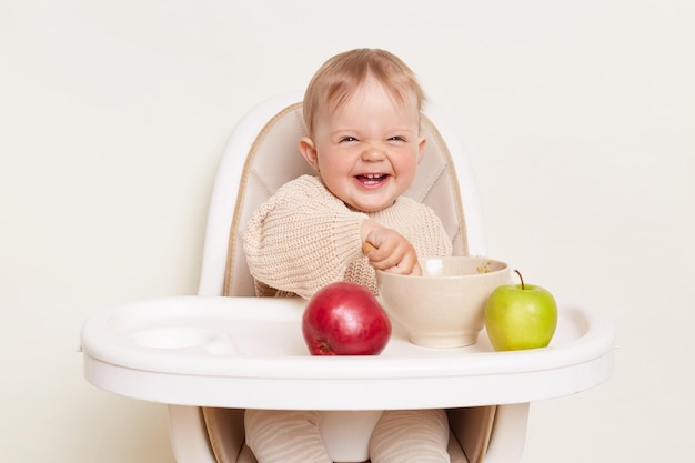 Plan Horizontal D'une Petite Fille Souriante Et Riante Portant Un Pull Beige Assis Sur Une Chaise Avec Une Expression Positive Mangeant Du Porridge Ou De La Purée De Fruits Prenant Son Petit Déjeuner