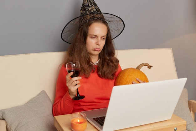 Plan horizontal d'une triste femme caucasienne portant un chapeau de sorcière travaillant sur un ordinateur portable à l'intérieur de la maison et buvant du vin dans un verre contrarié de travailler pendant Halloween