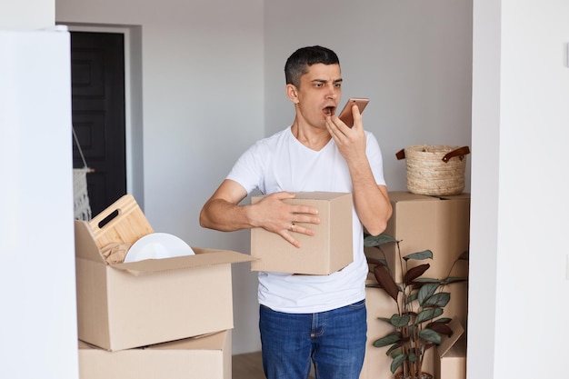 Plan intérieur d'un bel homme agressif en colère portant un t-shirt blanc debout avec une boîte en carton tenant un téléphone portable et criant de colère tout en enregistrant un message vocal