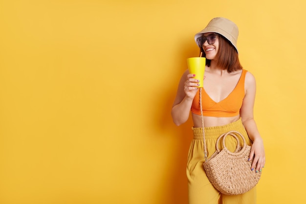 Plan intérieur d'une belle femme aux cheveux noirs portant des lunettes de soleil portant des vêtements d'été posant isolé sur fond jaune buvant un cocktail en regardant de côté l'espace de copie pour la publicité