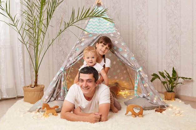 Plan intérieur d'une brune satisfaite d'un homme caucasien portant un t-shirt blanc allongé sur le sol avec ses petites filles près de sa tente peetee charmants enfants étreignant son père et exprimant des émotions positives