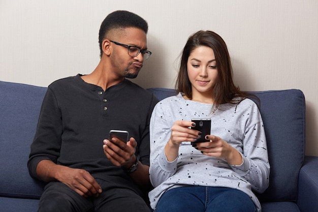 Plan intérieur d'un couple appréciant le contenu multimédia dans un téléphone intelligent assis sur un canapé confortable à la maison, tenant des téléphones mobiles