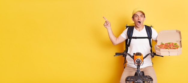 Plan intérieur d'un coursier étonné choqué à vélo tenant une boîte à pizza en carton ouverte et pointant vers l'espace de copie pour la publicité garde la bouche ouverte isolée sur fond jaune