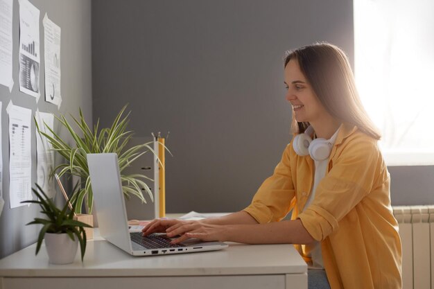 Plan intérieur d'une femme d'affaires portant une chemise jaune à l'aide d'un ordinateur portable et assise à la table du bureau avec une fenêtre en arrière-plan tapant sur l'écriture au clavier