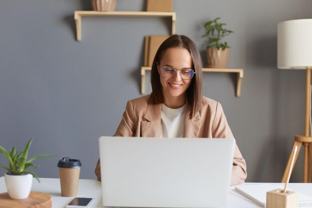 Plan intérieur d'une femme d'affaires positive souriante et heureuse aux cheveux bruns portant une veste beige travaillant en ligne sur un ordinateur portable en tapant sur un clavier ayant une expression positive