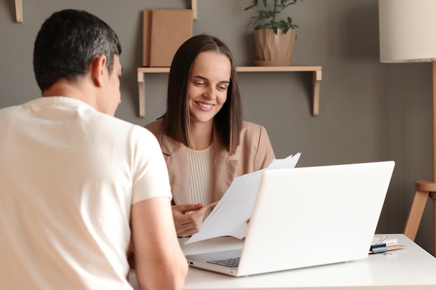 Plan intérieur d'une femme d'affaires souriante travaillant avec un nouveau partenaire montrant des documents assis à table au bureau devant des collègues portables négociant un nouveau contrat