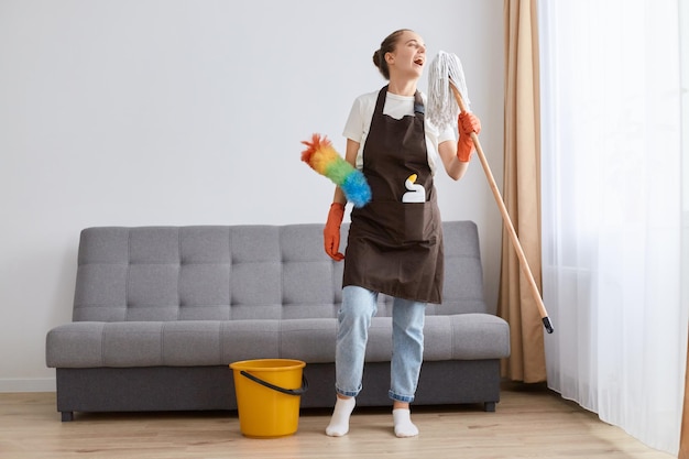 Plan intérieur d'une femme au foyer heureuse et positive nettoyant le sol avec une vadrouille à la maison en chantant et en s'amusant tout en nettoyant sa maison debout près d'un canapé et d'un seau jaune