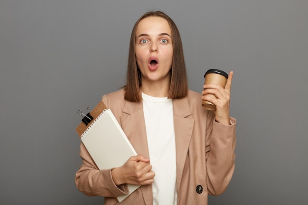 Plan intérieur d'une femme aux cheveux bruns caucasienne inspirée portant une veste tenant un presse-papiers avec des documents et un café à emporter pointant le doigt vers le haut ayant une idée posant isolé sur fond gris