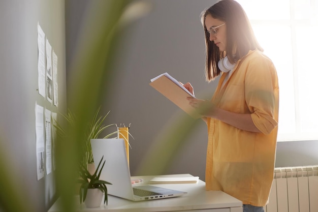 Plan intérieur d'une femme aux cheveux bruns portant une chemise jaune debout avec un organisateur près de son lieu de travail et vérifiant les informations femme secrétaire travaillant avec l'horaire du patron