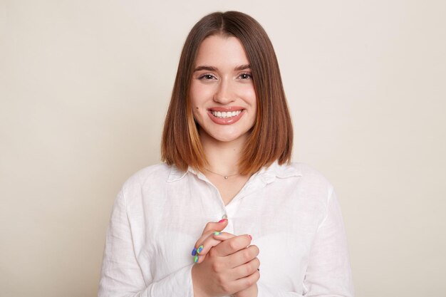 Plan intérieur d'une femme aux cheveux noirs optimiste positive portant une chemise posant isolé sur fond blanc regardant la caméra avec une expression positive garde les mains ensemble