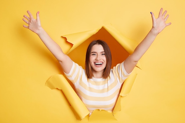 Plan intérieur d'une femme brune heureuse étonnée se tient dans un trou de papier déchiré posant avec les bras levés exprimant le bonheur en regardant à travers la percée du fond jaune