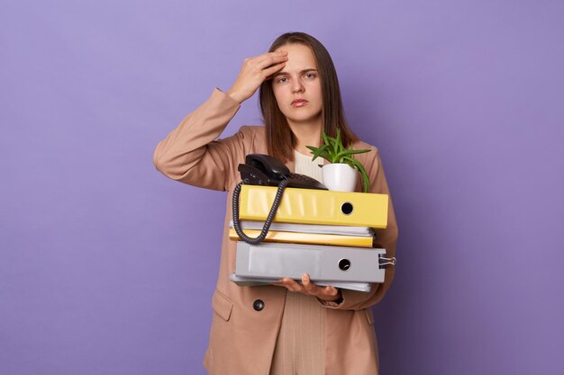 Plan intérieur d'une femme caucasienne triste et malsaine portant une veste de style officiel tenant des dossiers isolés sur fond violet ayant des maux de tête a beaucoup de travail