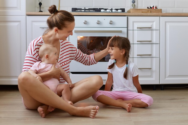 Plan intérieur d'une femme avec une coiffure en chignon portant une chemise rayée de style décontracté assise sur le sol dans la cuisine avec ses enfants exprimant des émotions positives en parlant et en riant