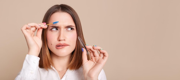 Plan intérieur d'une femme confuse perplexe en chemise blanche debout avec une brosse à sourcils et une pince à épiler dans les mains regardant loin l'espace de copie pour la publicité ou le texte promotionnel