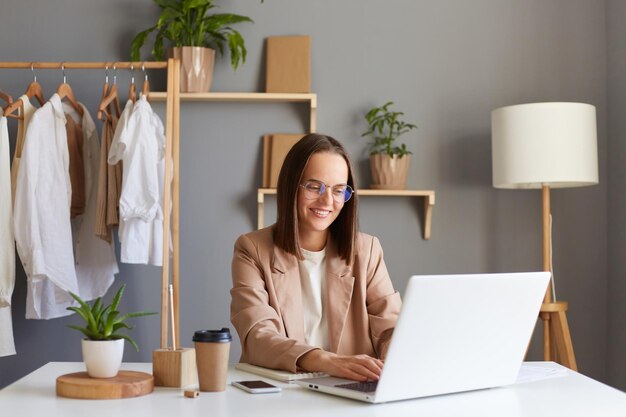 Plan intérieur d'une femme designer positive souriante dans des verres travaillant sur un ordinateur portable dans le salon contre des vêtements accrochés sur une étagère styliste féminine portant une veste beige tapant sur un clavier d'ordinateur portable