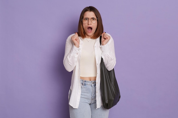 Plan intérieur d'une femme effrayée effrayée portant des vêtements élégants tenant un sac posant isolé sur fond violet debout avec les poings serrés étant désespéré en criant