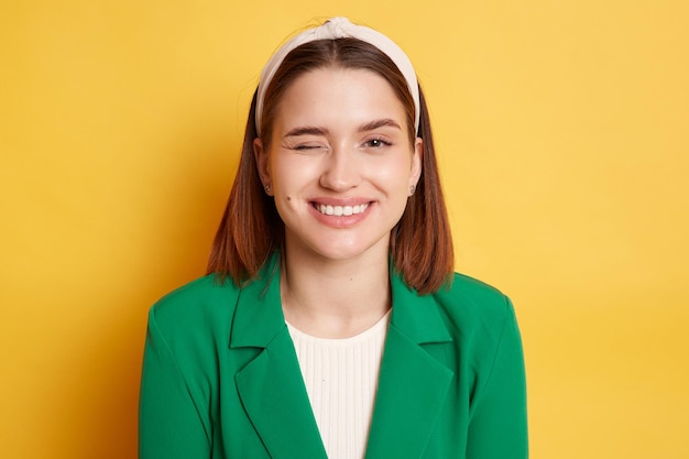 Plan intérieur d'une femme enfantine ludique portant une veste verte posant isolée sur fond jaune regardant la caméra avec un sourire à pleines dents et un clin d'œil flirtant avec son petit ami