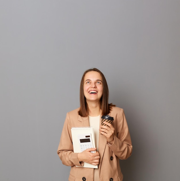 Plan intérieur d'une femme excitée étonnée aux cheveux bruns portant une veste beige debout contre un mur gris tenant des papiers et du café pour aller chercher et copier l'espace pour la publicité