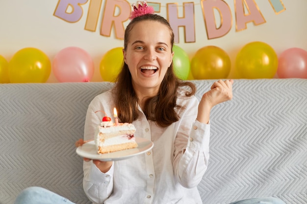 Plan intérieur d'une femme excitée extrêmement heureuse portant une chemise blanche assise sur la toux, tenant un gâteau avec une bougie, un poing fermé, une décoration festive sur fond.