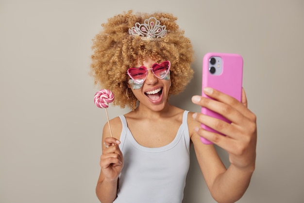 Plan intérieur d'une femme gaie aux cheveux bouclés et touffus portant une couronne et un t-shirt décontracté lunettes de soleil coeur rose applique des patchs de beauté sous les yeux pour éliminer les poches prend un selfie tient une sucette sur un bâton