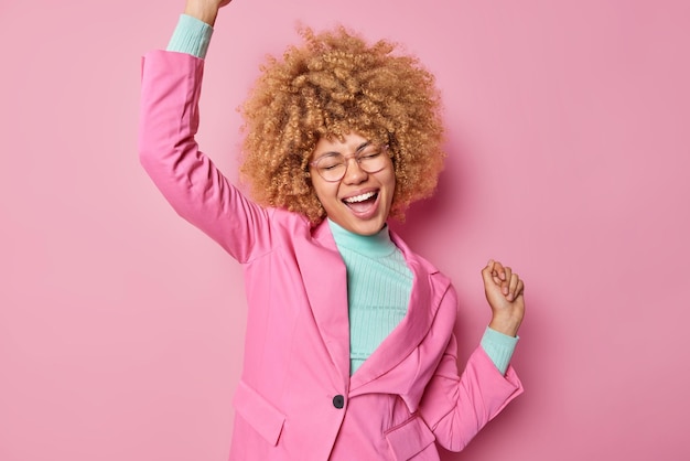 Plan intérieur d'une femme heureuse et optimiste aux cheveux bouclés secoue les bras pour faire triompher la danse s'exclame bruyamment porte des lunettes veste formelle isolée sur fond rose Une femme d'affaires prospère ressent de la joie