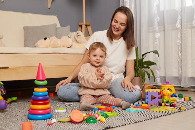 Plan intérieur d'une femme heureuse et de sa jolie fille assise sur le sol et passant du temps ensemble enfant applaudissant la famille posant parmi de nombreux jouets colorés