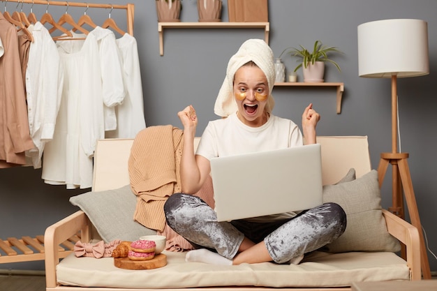 Plan intérieur d'une femme joyeuse et ravie portant un pantalon gris T-shirt blanc et avec une serviette sur la tête assise sur la toux avec un ordinateur portable regardant l'écran et les poings serrés se réjouissant de parier et de gagner