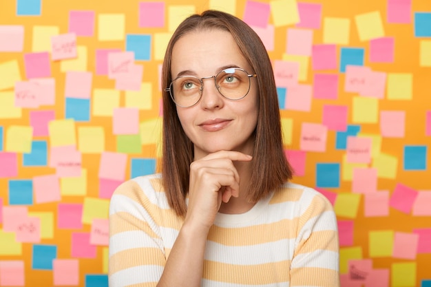 Plan intérieur d'une femme pensive et réfléchie aux cheveux bruns en t-shirt rayé debout contre un mur jaune avec des autocollants colorés tenant son menton en détournant les yeux en rêvant