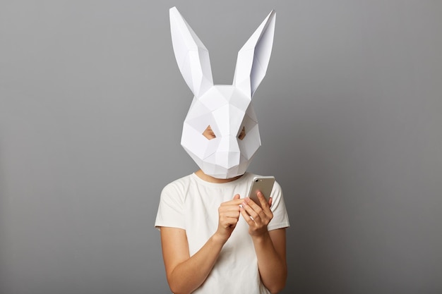 Plan intérieur d'une femme portant un masque de lapin et un sweat-shirt orange posant isolé sur fond gris tenant un smartphone dans les mains à l'aide d'un téléphone mobile naviguant sur Internet