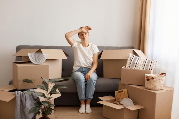 Plan Intérieur D'une Femme Portant Un T-shirt Blanc Et Un Jean Assise Sur Un Canapé Lors D'un Déménagement Dans Un Nouvel Endroit En Gardant La Main Sur Son Front Fatiguée Et épuisée