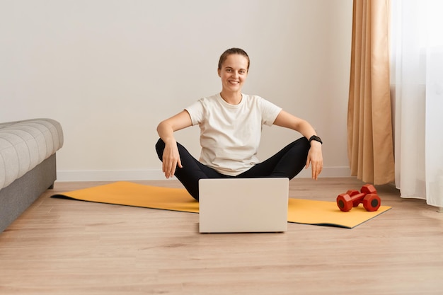 Plan intérieur d'une femme portant un t-shirt blanc et des leggins noirs assis sur un tapis avec les bras croisés posant devant un ordinateur portable en regardant une vidéo de tutoriel pour la pratique du yoga