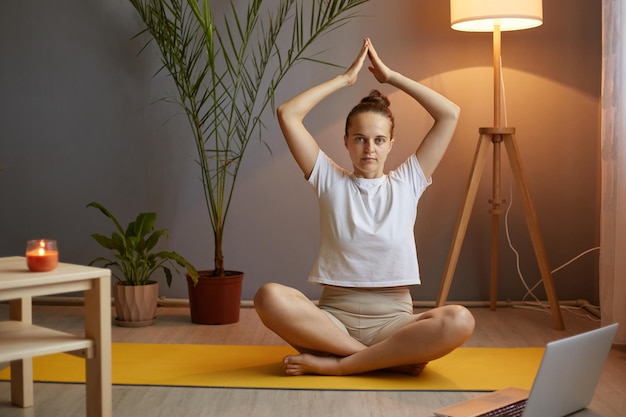 Plan intérieur d'une femme portant un t-shirt blanc pratiquant le yoga et étirant le corps les bras levés garde les paumes ensemble en regardant l'ordinateur portable de la caméra pour le cours en ligne