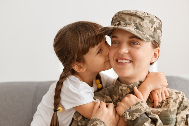 Plan intérieur d'une femme soldat heureuse souriante portant un uniforme de camouflage et une casquette posant avec sa fille mère rentre de la guerre enfant manquant tellement étreindre et embrasser maman