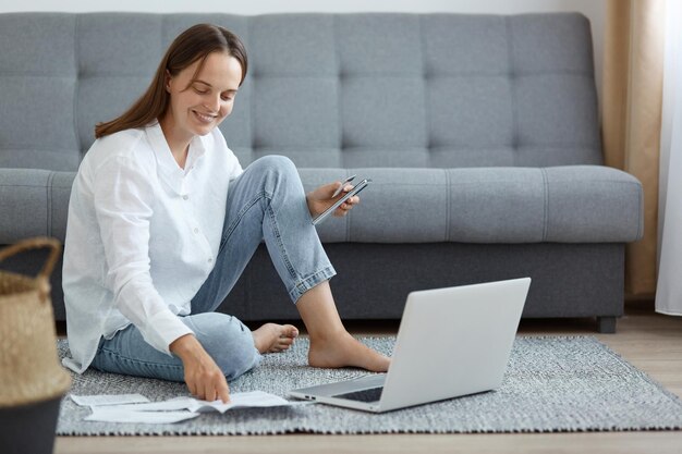 Plan intérieur d'une femme souriante portant une chemise blanche et un jean assis sur le sol près d'un canapé à l'aide d'un ordinateur portable tenant une carte de crédit vérifiant les informations sur les factures de services publics sur papier