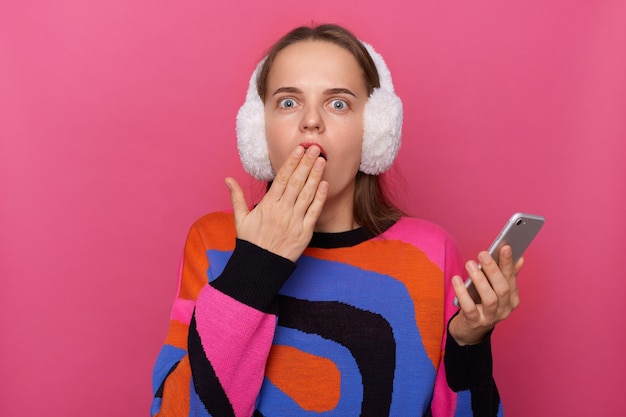 Plan intérieur d'une femme surprise étonnée, choquée, portant un pull chaud et des cache-oreilles en fourrure, debout, isolée sur fond rose, lisant les dernières nouvelles sur les réseaux sociaux