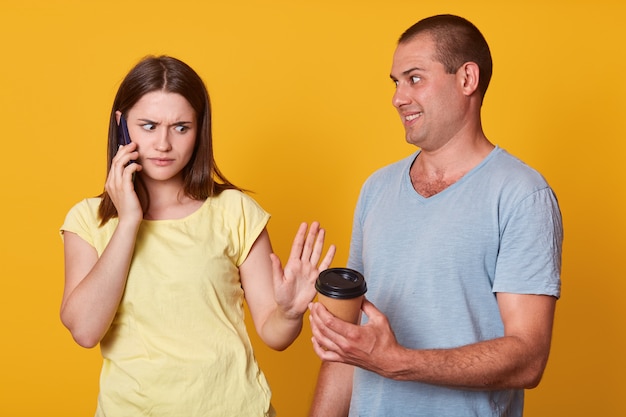 Plan intérieur d'une femme triste regarde avec une expression bouleversée au gars offre son café à emporter, a une expression faciale bouleversée, parle par téléphone portable, debout contre le mur jaune du studio, les mariés portent avec désinvolture.